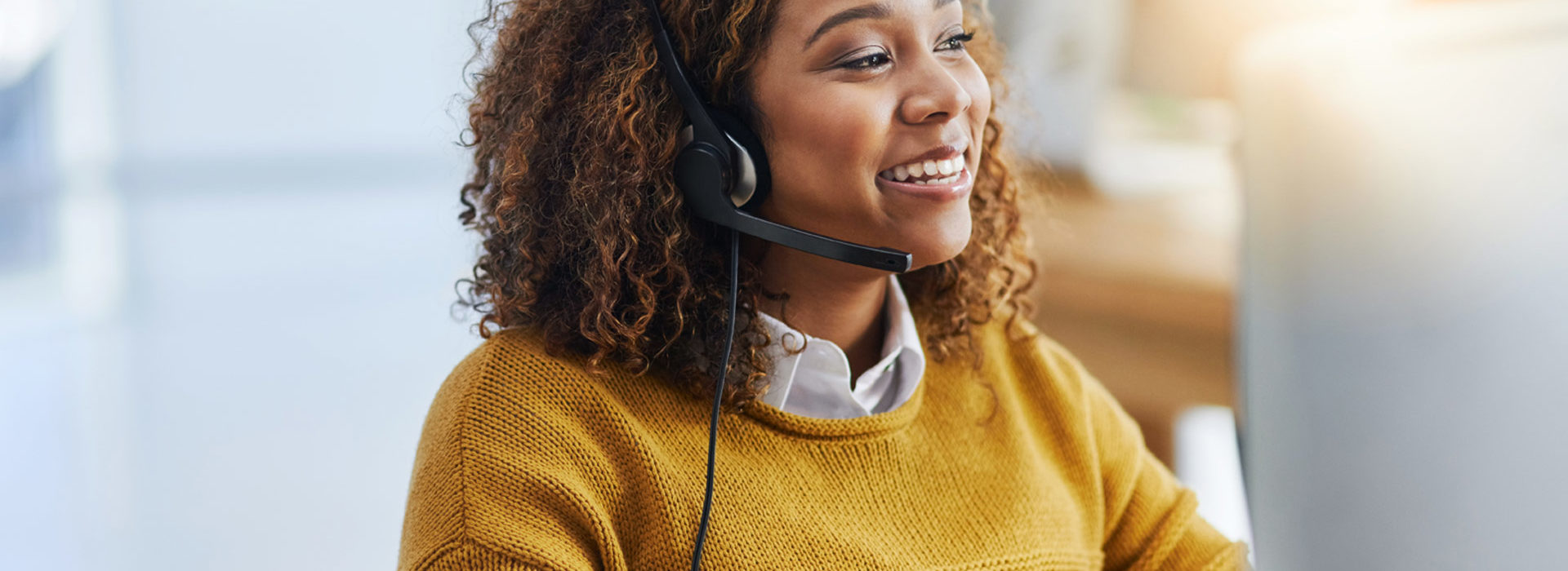 Une femme souriante au téléphone avec un casque devant son ordinateur portant un pull jaune - assistance aux personnes