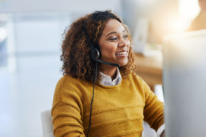 Une femme souriante au téléphone avec un casque devant son ordinateur portant un pull jaune