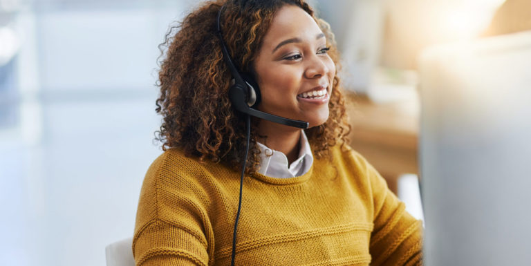 Une femme souriante au téléphone avec un casque devant son ordinateur portant un pull jaune - assistance aux personnes