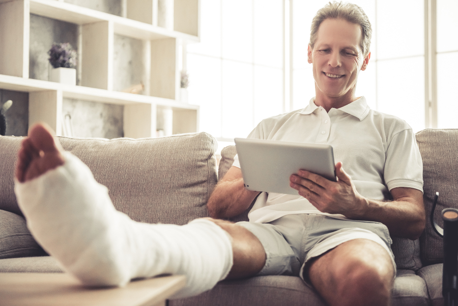 Un homme en arrêt de travail avec une jambe cassée regardant sa tablette dans son canapé