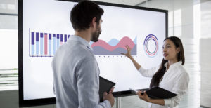 Male and female colleague standing by large screen in meeting room, going over financial presentation shown on screen in meeting room.