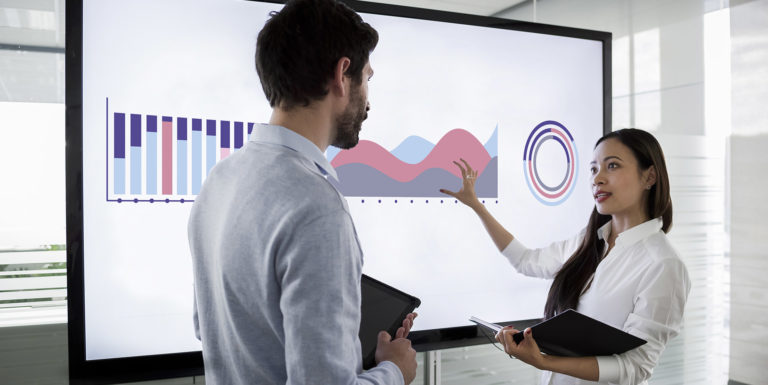 Male and female colleague standing by large screen in meeting room, going over financial presentation shown on screen in meeting room.