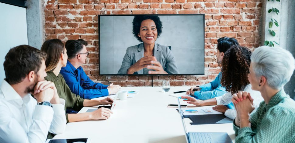 Des hommes et des femmes en réunion au bureau devant un écran projetant une femme en viso conférence - ressources humaines entreprises