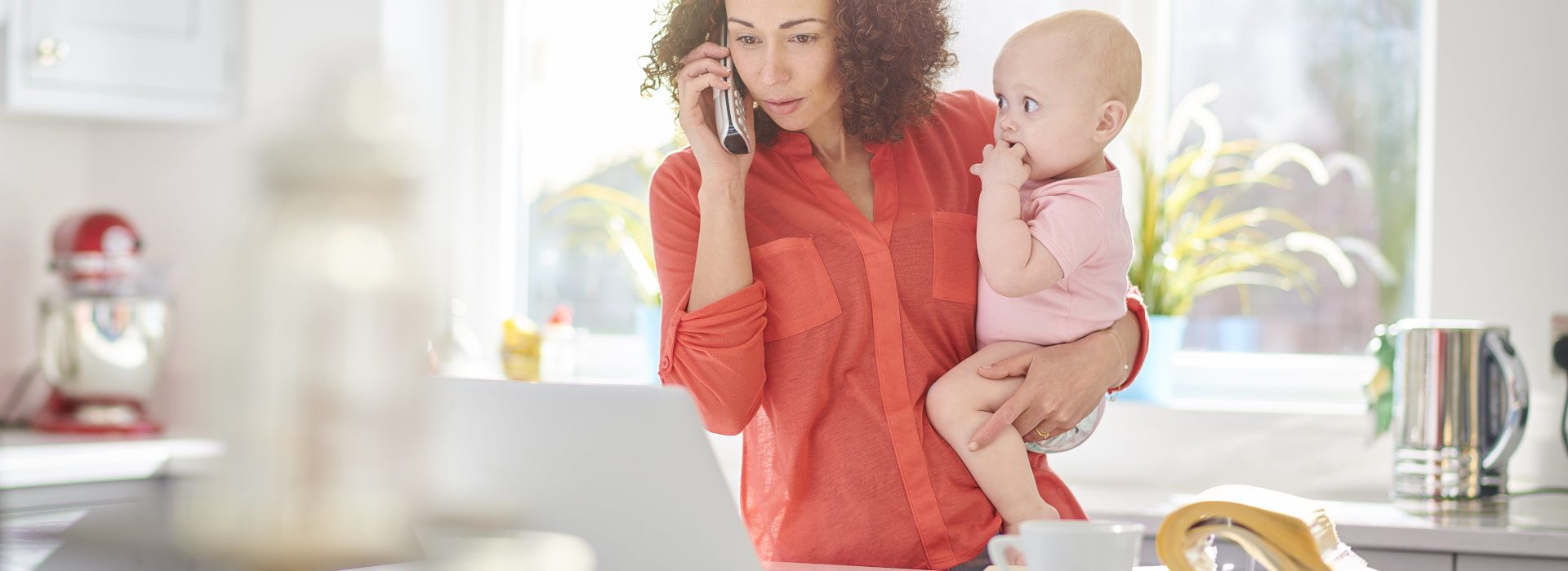 Femme à l’étranger tenant son bébé dans les bras tout en téléphonant devant son ordinateur au sujet de sa protection sociale grâce la caisse des Français à l’étranger