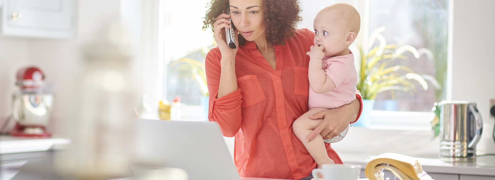 Une femme tenant son bébé dans les bras et téléphonant devant son ordinateur au sujet de sa protection sociale grâce la caisse des Français à l’étranger Femme à l’étranger tenant son bébé dans les bras tout en téléphonant devant son ordinateur au sujet de sa protection sociale grâce la caisse des Français à l’étranger