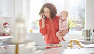 Femme à l’étranger tenant son bébé dans les bras tout en téléphonant devant son ordinateur au sujet de sa protection sociale grâce la caisse des Français à l’étranger