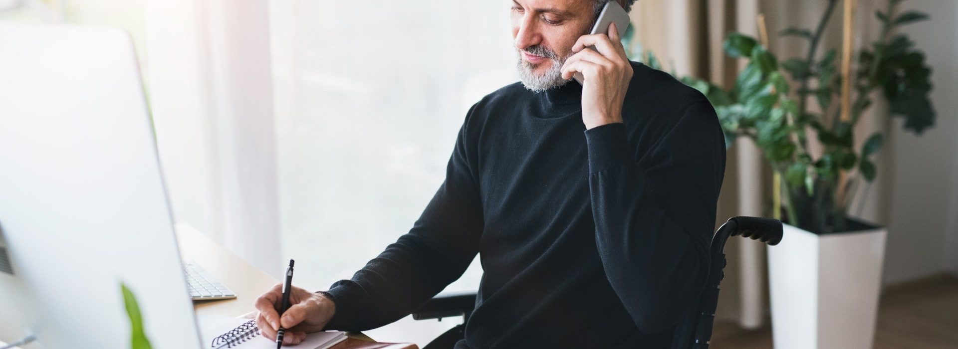 Un homme en fauteuil roulant à son bureau, prenant des notes et étant au téléphone sur l'espace client entreprise épargne salariale