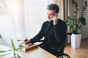Un homme en fauteuil roulant à son bureau, prenant des notes et étant au téléphone sur l'espace client entreprise épargne salariale