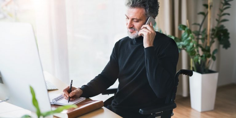 Un homme en fauteuil roulant à son bureau, prenant des notes et étant au téléphone sur l'espace client entreprise épargne salariale